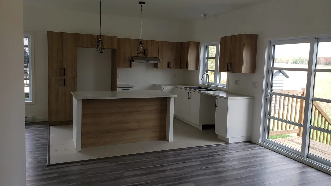 Cuisine d’un logement de duplex neuf : armoires de bois, îlot et porte-fenêtre sur le balcon