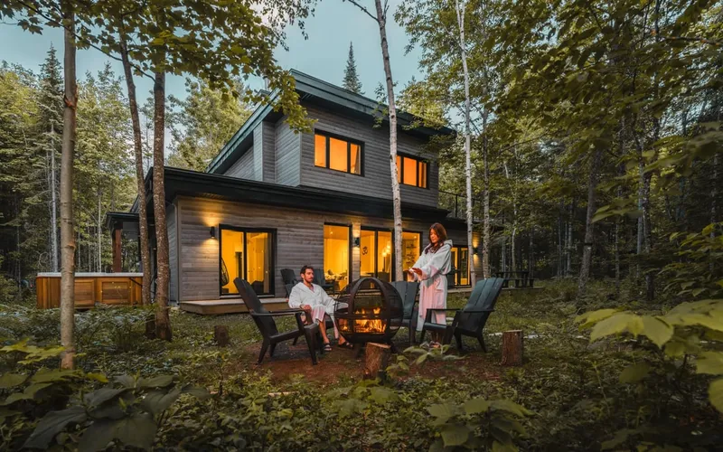 Chalet locatif haut de gamme en bois au crépuscule, avec foyer extérieur et spa nordique en forêt