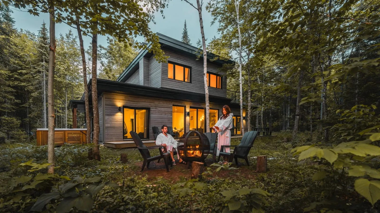 Chalet locatif haut de gamme en bois au crépuscule, avec foyer extérieur et spa nordique en forêt