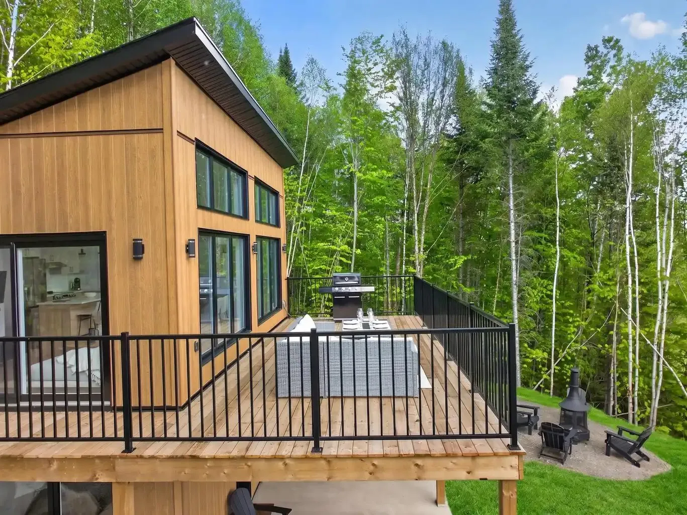 Terrasse de bois à l'étage avec garde-corps noir, barbecue et mobilier de rotin, ouverte sur la forêt
