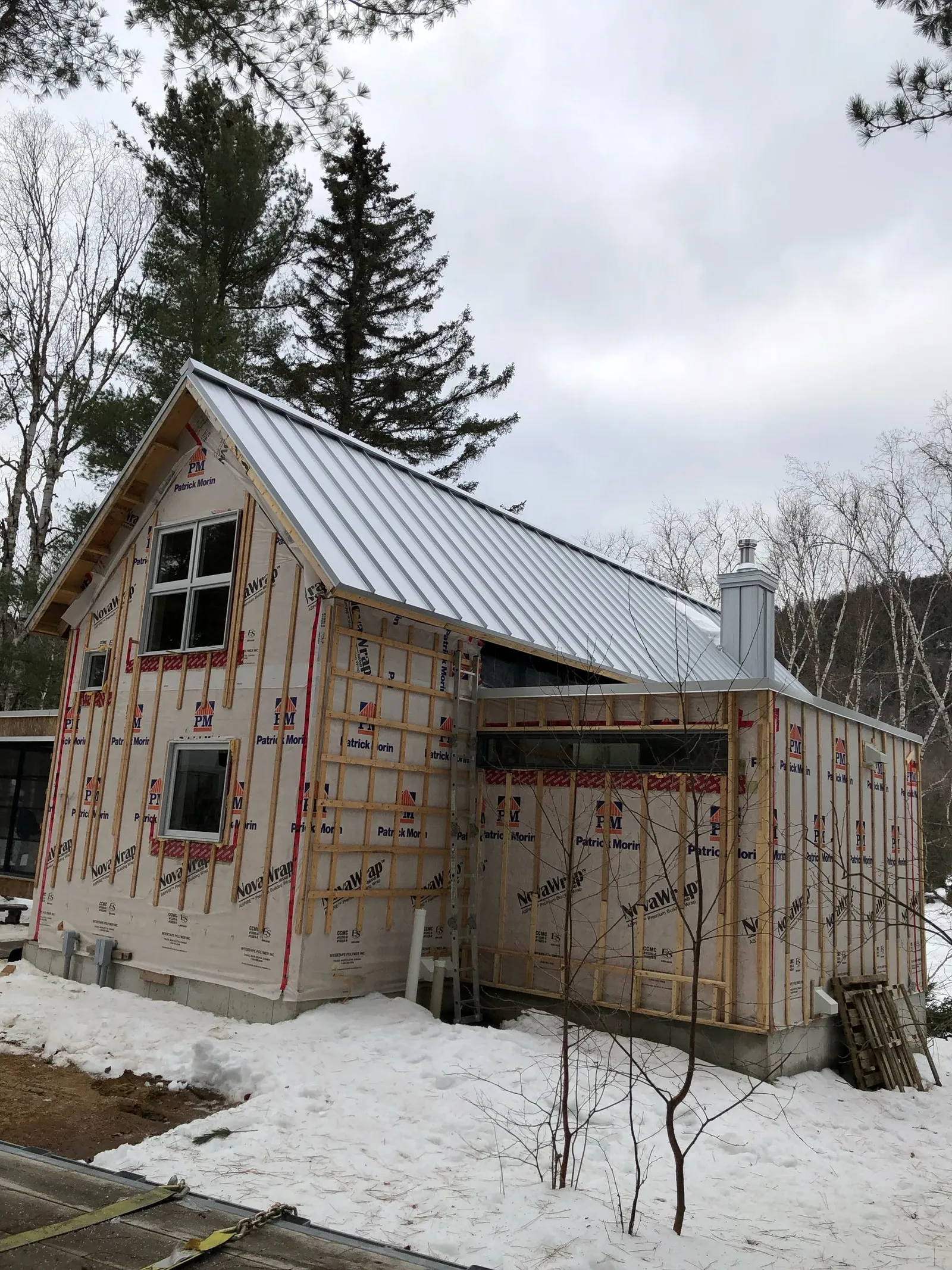 Le chalet en phase d’enveloppe, pignon et toiture métallique, vu du terrain boisé enneigé