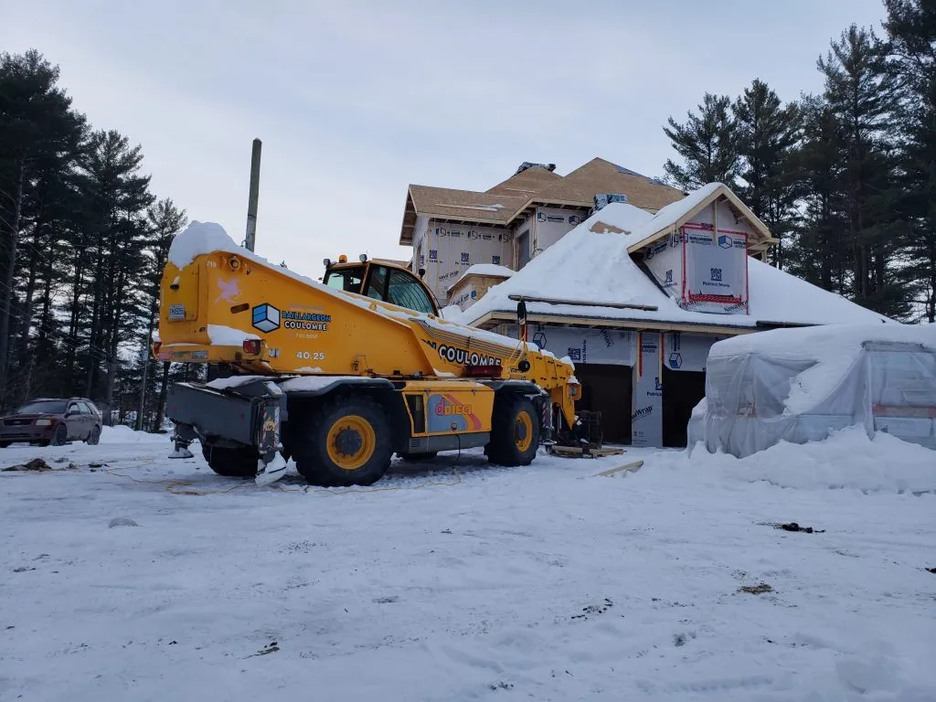 Chariot télescopique couvert de neige devant le bâtiment en chantier, matériaux protégés sous une bâche et bancs de neige
