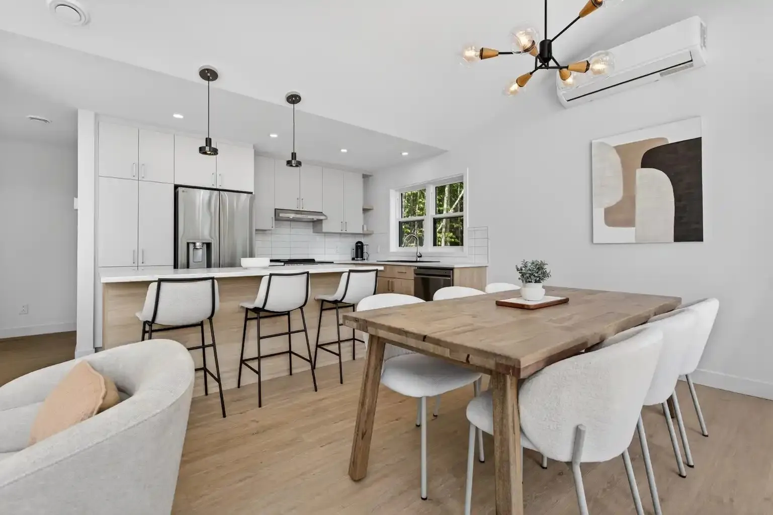 Salle à manger avec table de bois, chaises blanches et suspension noire, ouverte sur la cuisine